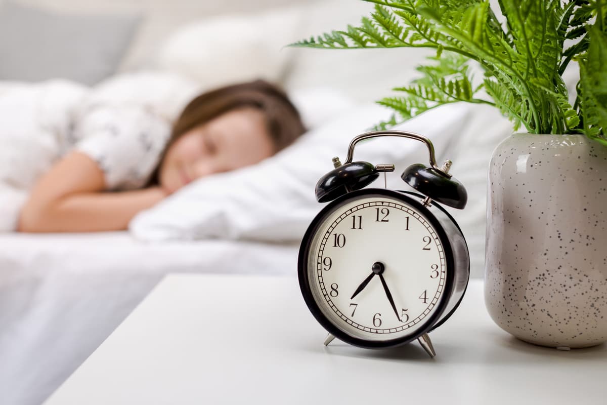 A young girl asleep in a bright bedroom beside an alarm clock